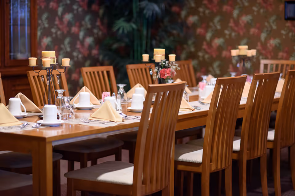 A long wooden dining table set with beige folded napkins, white cups, glasses, silverware, and candle holders with multiple candles. The background features floral wallpaper and wooden chairs arranged around the table.