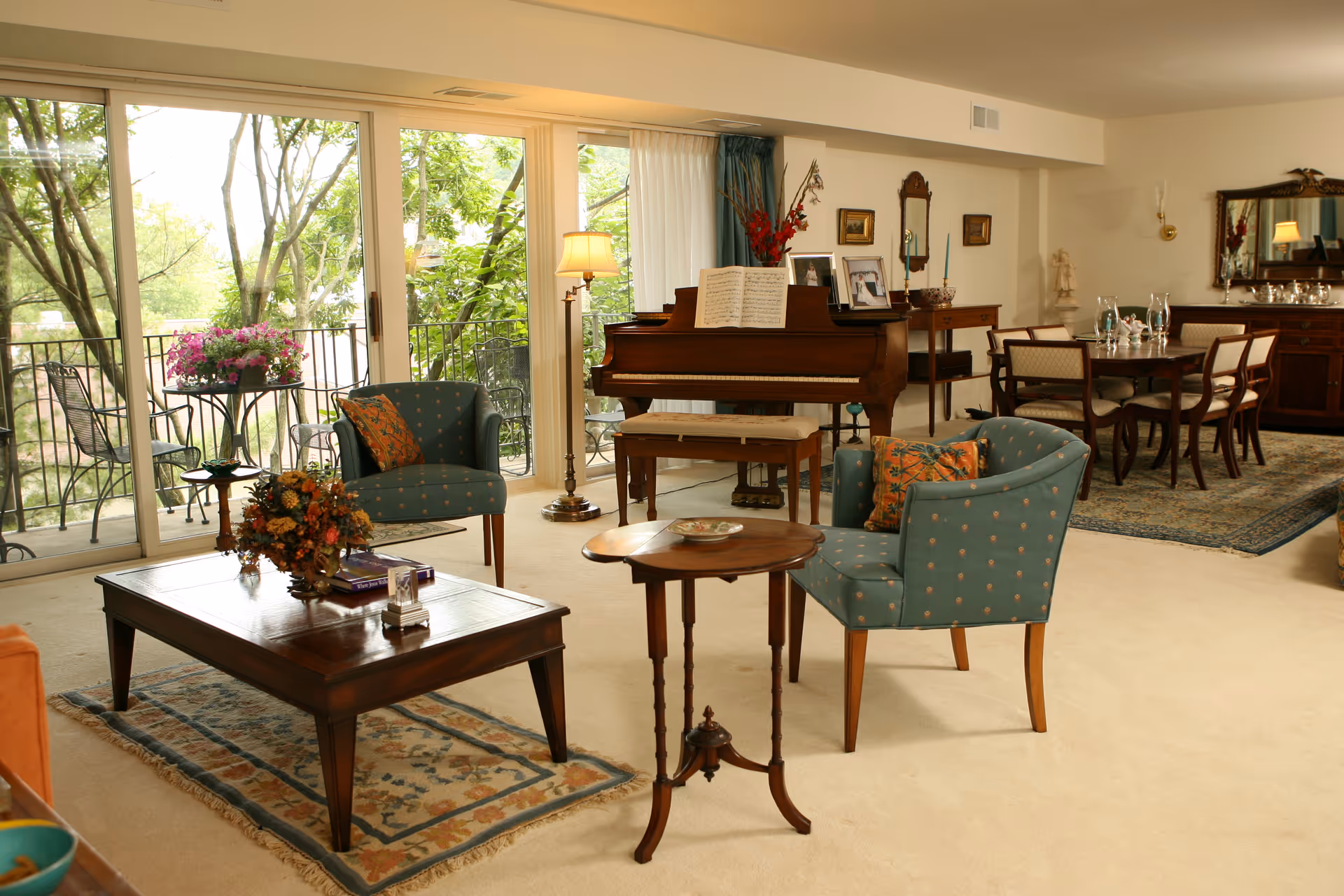 A spacious living room with large sliding glass doors opening to a balcony with outdoor seating and flowers. Inside, there are two blue upholstered chairs with patterned cushions, a wooden coffee table with a floral arrangement and books, a small round side table, and a grand piano with sheet music and framed photos on top. In the background, there is a dining area with a wooden dining table and chairs, a sideboard with a mirror, and decorative items including a statue and candlesticks.