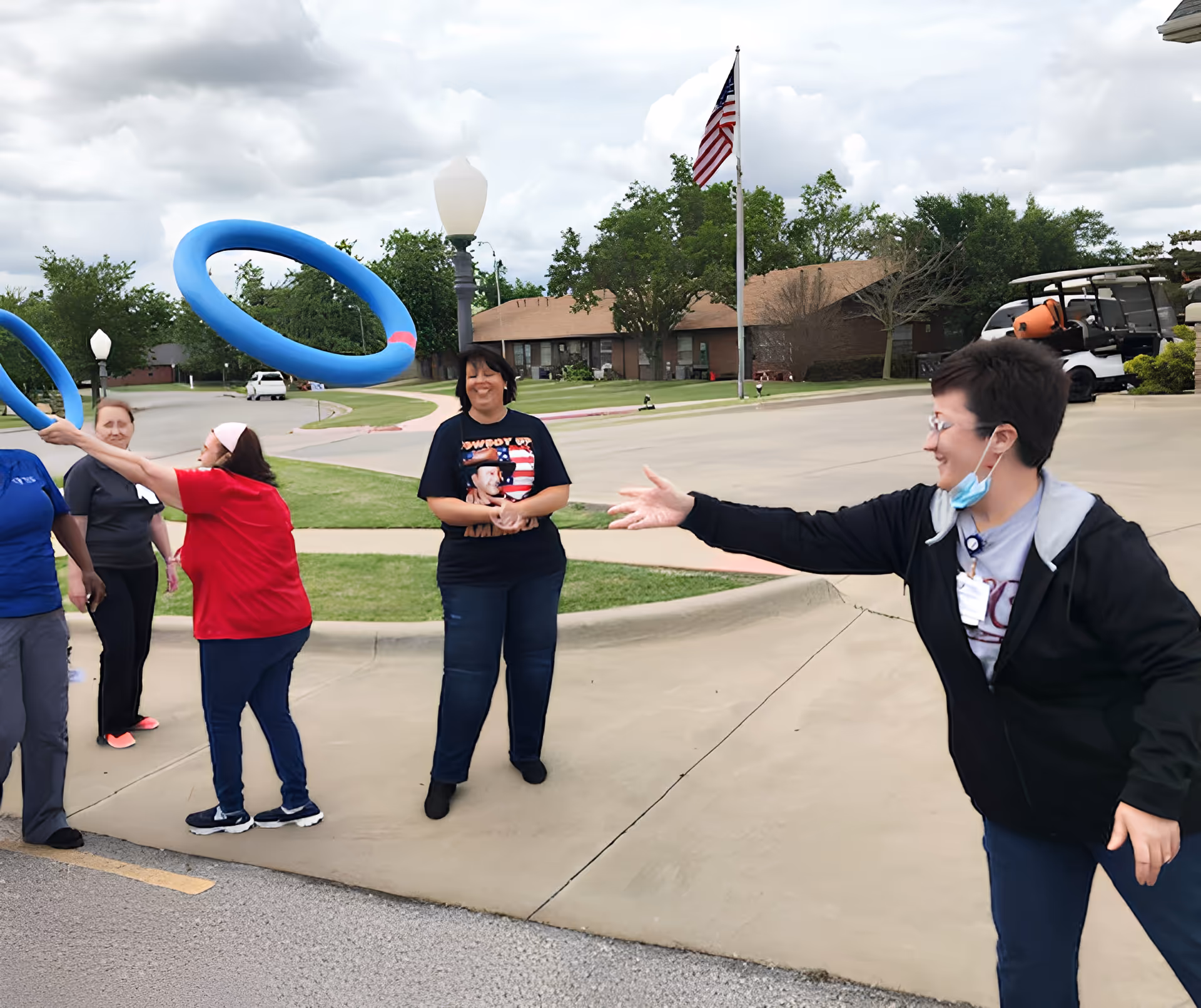 A group of women outdoors playing a ring toss game with large blue rings. One woman is actively throwing a ring while others watch and smile. The setting is a paved area with grass, trees, and buildings in the background, along with an American flag on a flagpole.