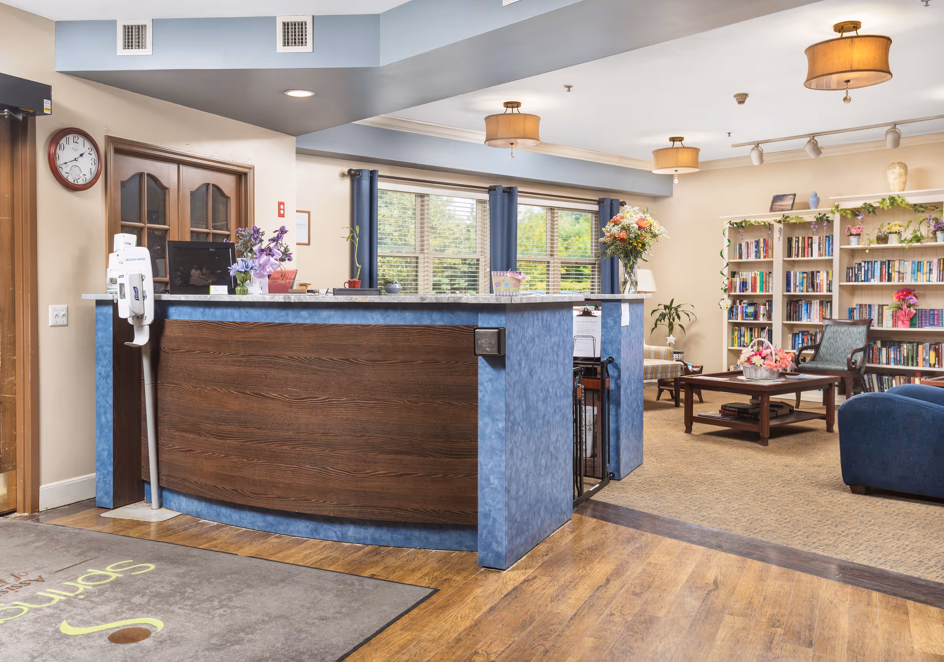 Reception area of Spring Oak at Forked River featuring a wooden and blue front desk with flowers and a computer on top. Behind the desk are windows with blue curtains letting in natural light. To the right, there is a cozy seating area with a coffee table, chairs, a bookshelf filled with books, and decorative plants and flowers. The floor is a combination of wood and carpet, and the ceiling has several light fixtures.
