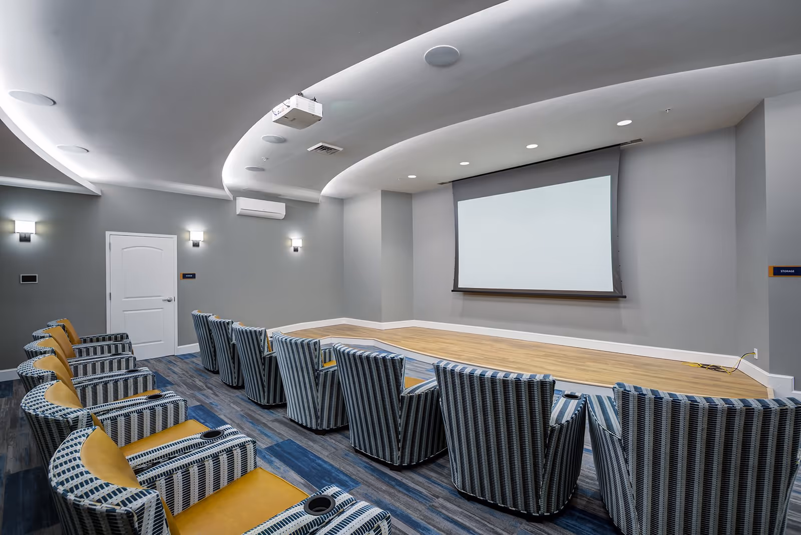 A modern small theater room with two rows of striped armchairs facing a large white projection screen on a gray wall. The room has a curved ceiling with recessed lighting and a projector mounted above. The floor is carpeted with a blue and gray pattern, and there is a wooden stage area in front of the screen.