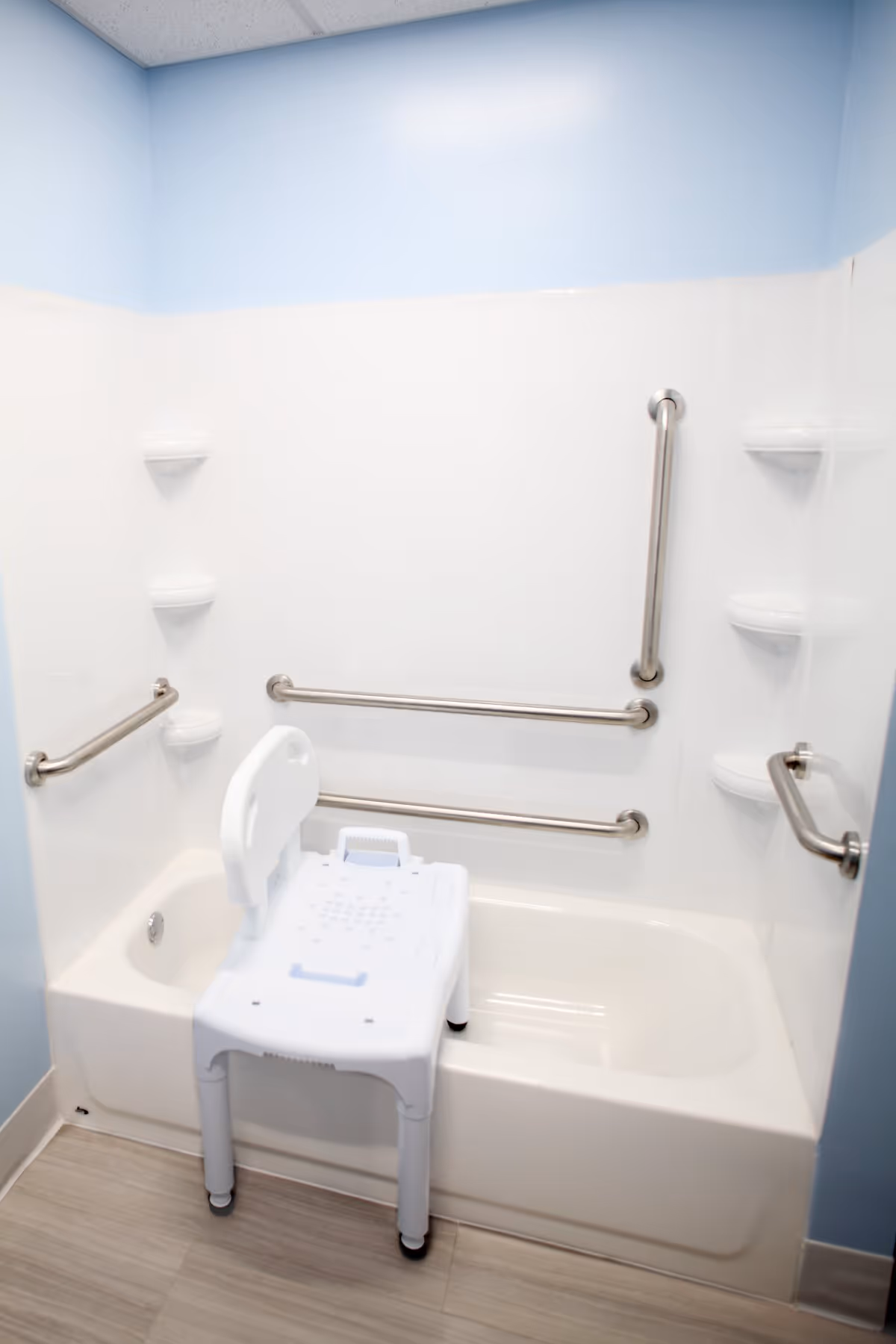 A bathtub with multiple stainless steel grab bars on the walls and a white plastic shower chair placed inside the tub. The walls are light blue and white, and the floor has light-colored tiles.