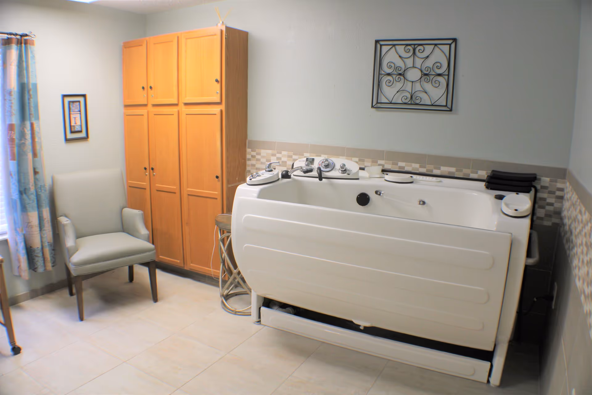 A room with a white walk-in bathtub equipped with control knobs and a handheld shower. Next to the bathtub is a wooden cabinet with multiple doors, a small round side table, and a gray upholstered armchair. The walls are light-colored with a decorative tile border and a metal wall art piece above the bathtub. A window with patterned curtains is partially visible on the left.