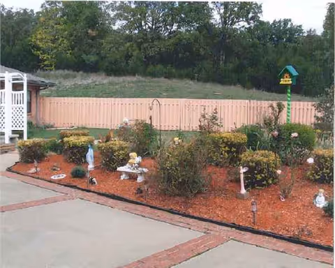 A well-kept outdoor garden with mulch, shrubs, statues, a birdhouse, and a peach fence.