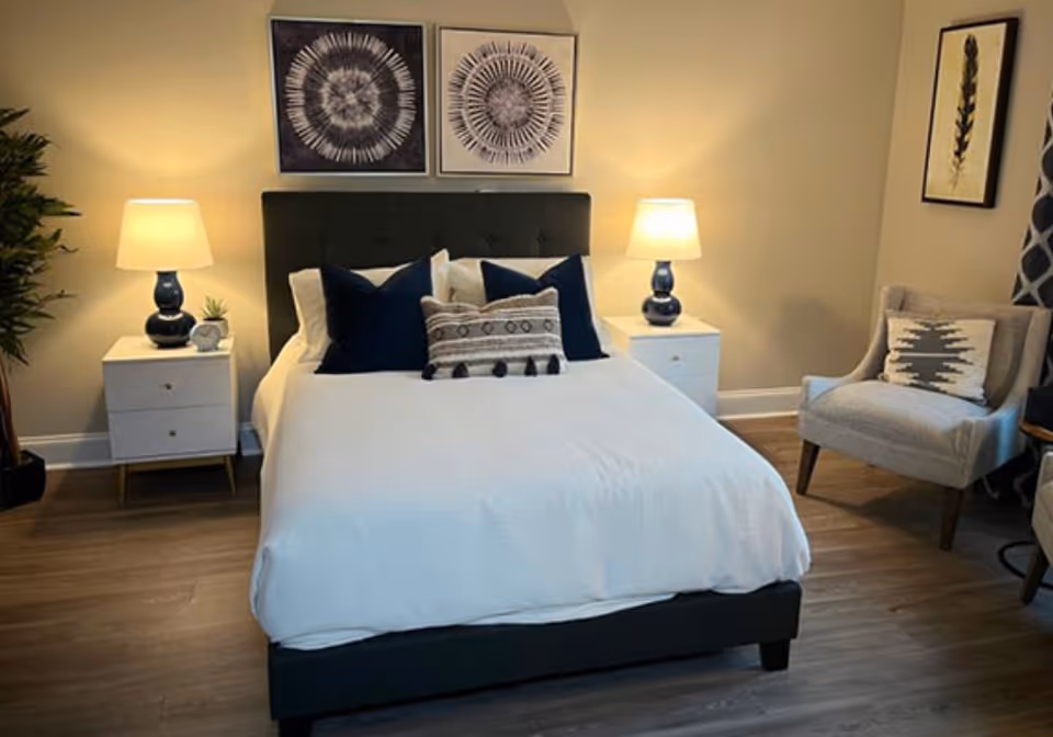 A neatly made bed with white bedding and decorative pillows in a bedroom. The bed has a dark upholstered headboard and is flanked by two white nightstands, each with a blue lamp. There are two framed circular abstract artworks above the bed. To the right, there is a gray armchair with a patterned pillow, and a framed botanical print on the wall. The floor is wooden, and there is a potted plant on the left side of the room.