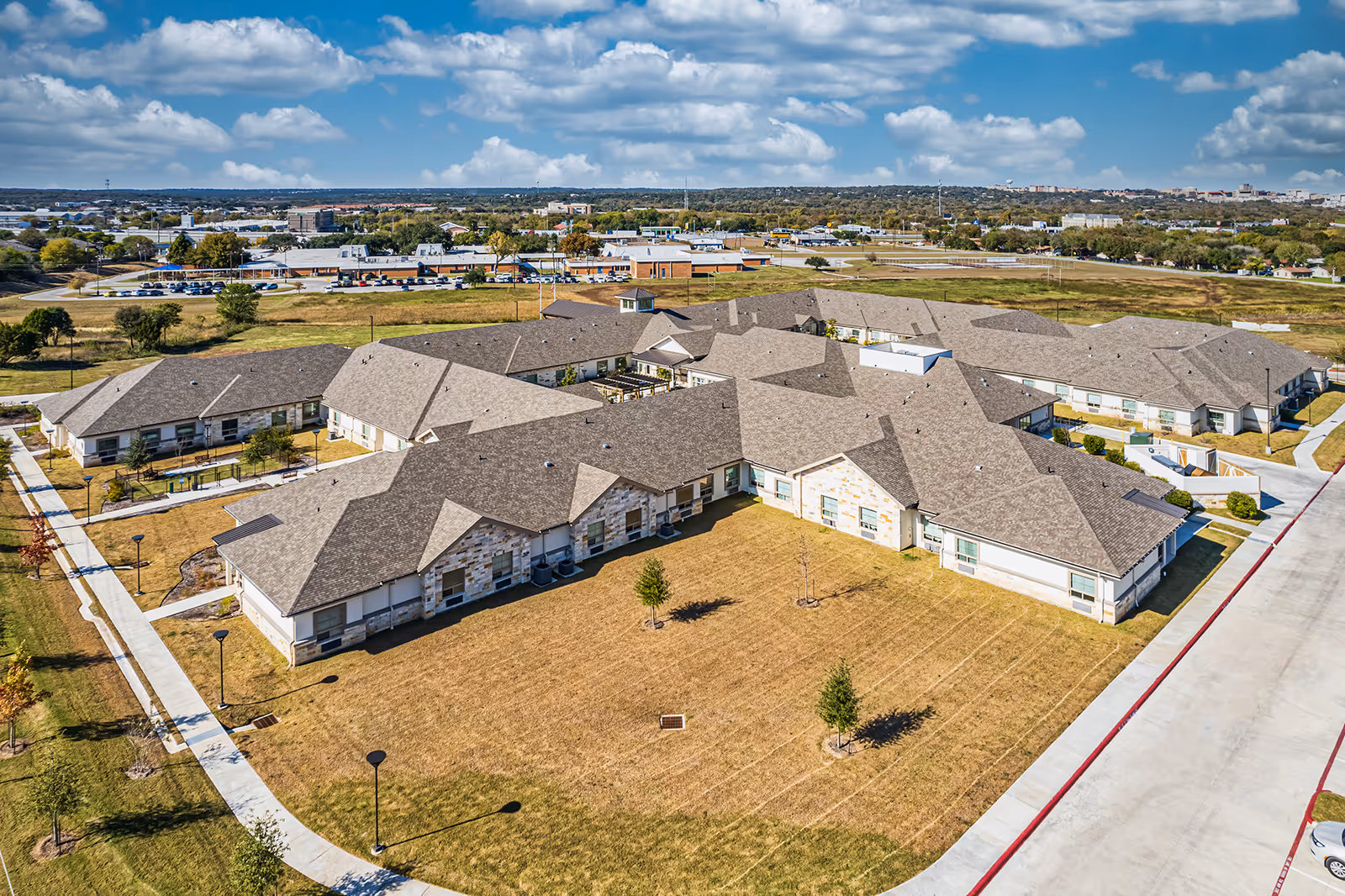 Aerial view of a single-story senior living campus with multiple connected buildings, lawns, walkways, and surrounding roads.