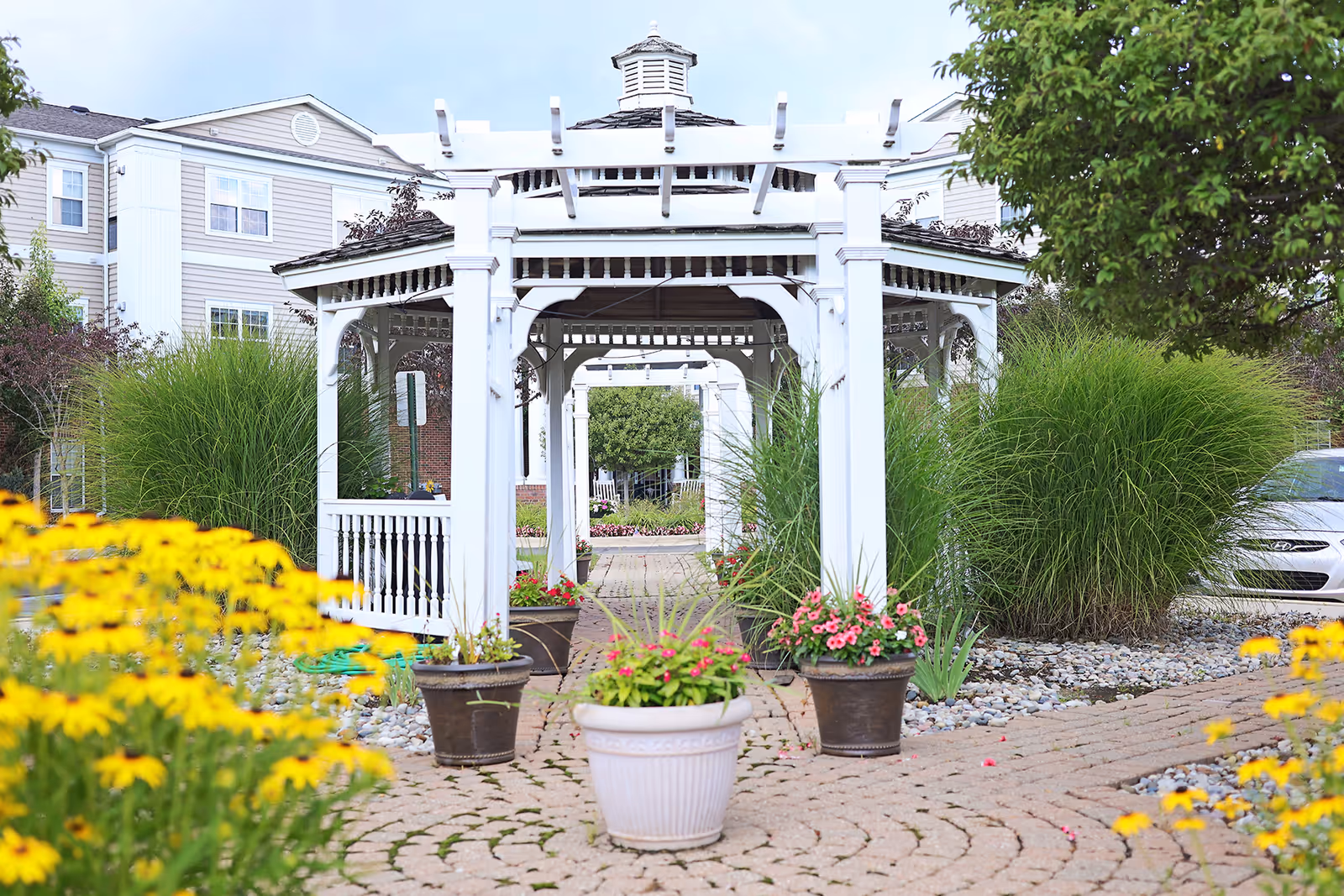A white wooden gazebo surrounded by potted flowers and lush greenery in a garden area with a paved walkway. In the background, there is a multi-story building and a parked car partially visible behind the bushes.