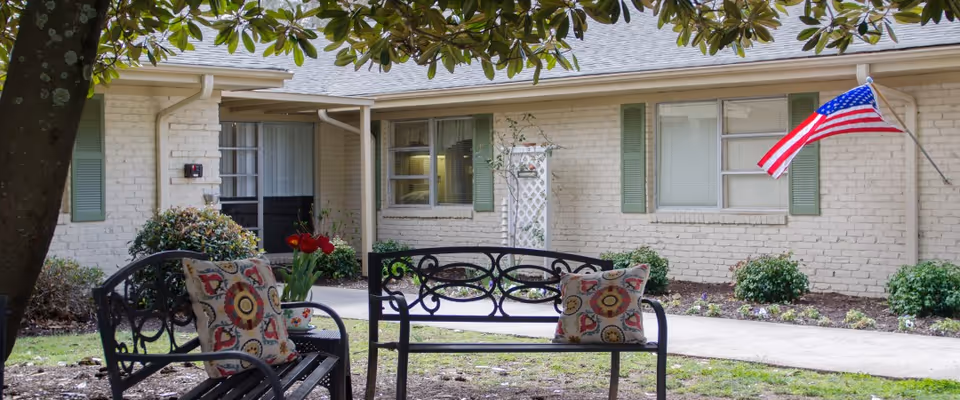 Front exterior of a single-story painted-brick building with an outdoor bench topped with decorative pillows and flowers, and an American flag.