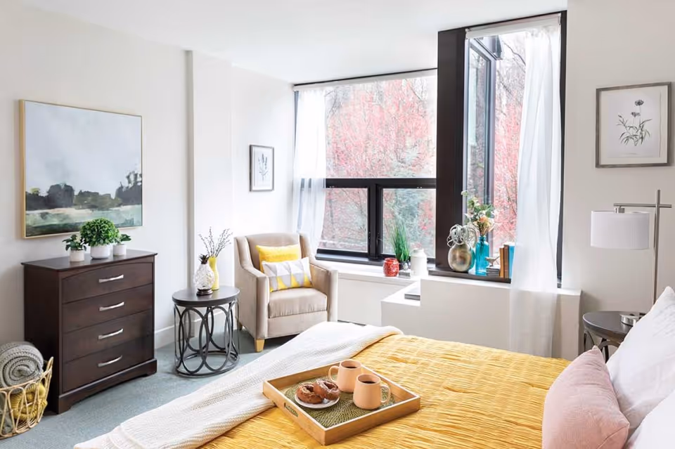 Sunlit bedroom with a yellow bedspread, a tray with mugs on the bed, an armchair by a large window, and a dark wood dresser.