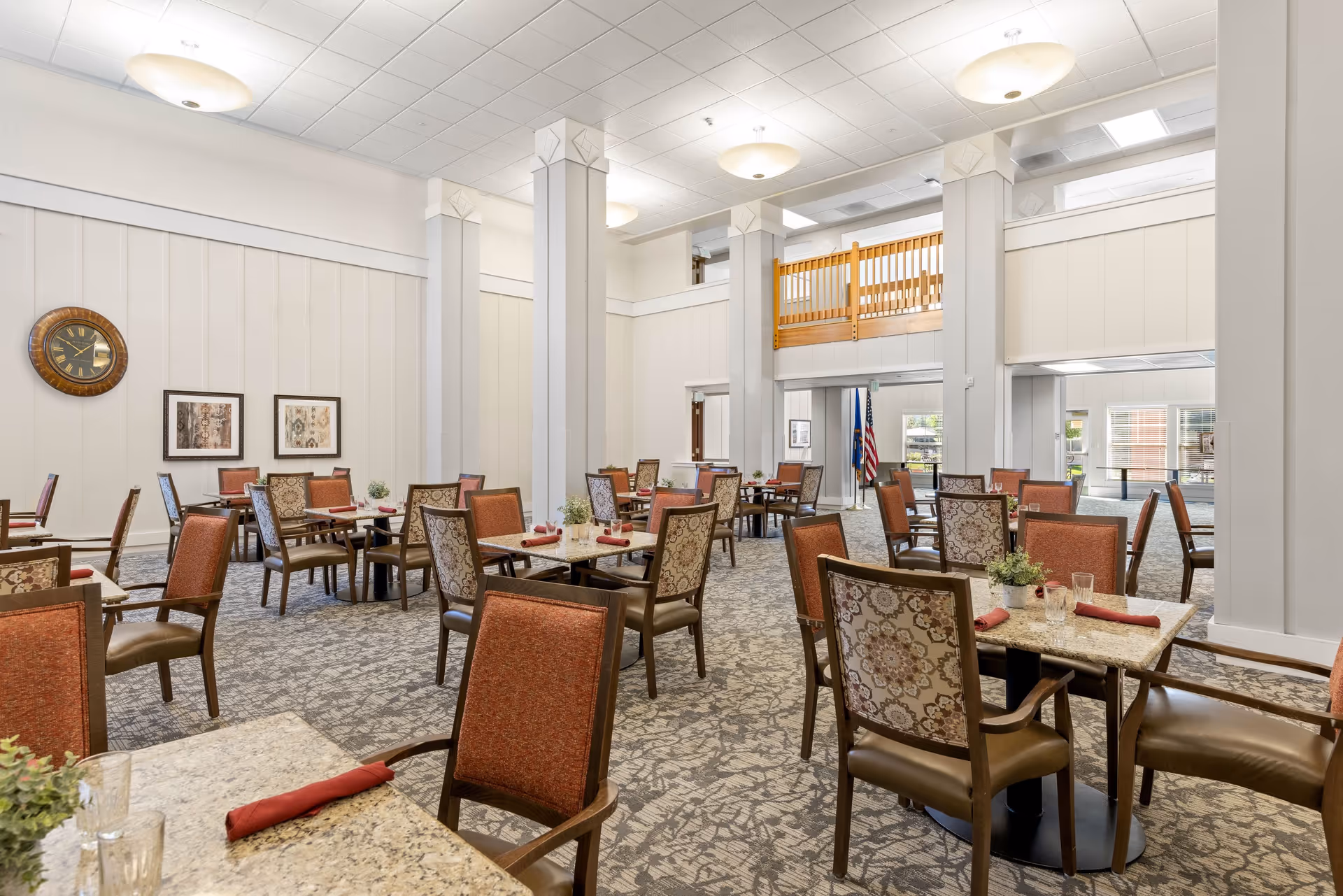 A spacious dining room with multiple tables and chairs arranged neatly. Each table has a granite top with a red napkin and glassware. The room features high ceilings with large round light fixtures, a patterned carpet, and light-colored walls adorned with framed artwork and a large clock. There is a wooden railing balcony visible on the upper level and an American flag near the entrance.