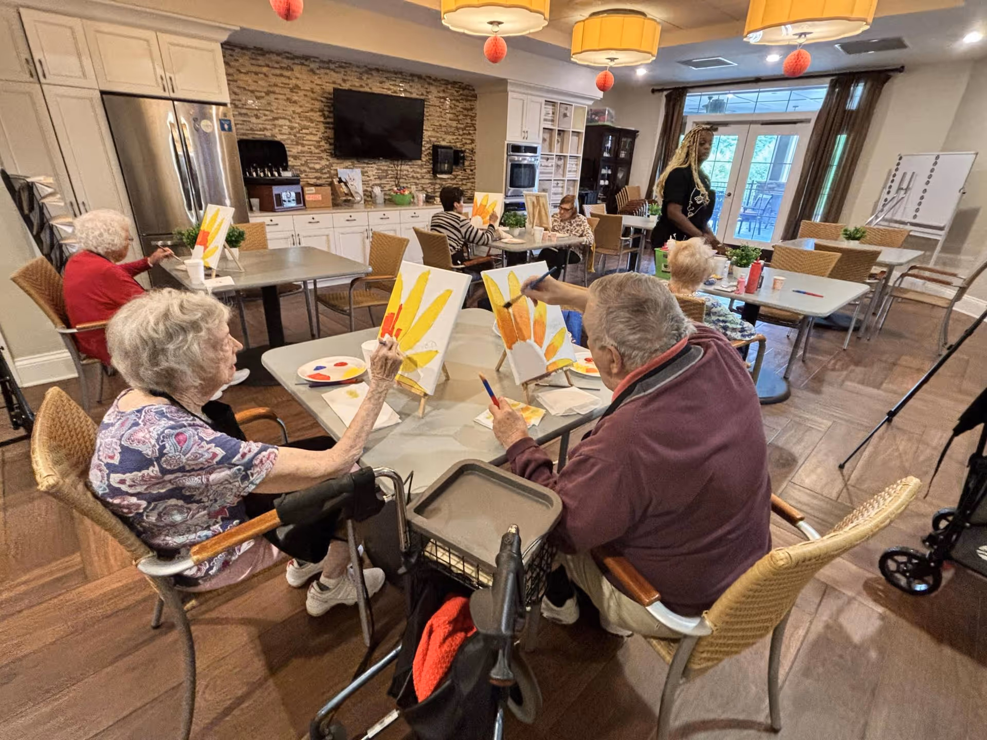 Several elderly individuals seated around tables in a well-lit room, engaged in painting on canvases. The room has a kitchen area with white cabinets, a stone backsplash, and a large TV mounted on the wall. There are large windows with curtains and hanging light fixtures above the tables. A caregiver or instructor is standing nearby assisting the group.