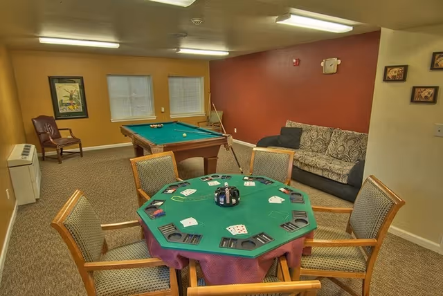 A cozy game room with a green octagonal card table surrounded by six chairs, playing cards and card holders on the table, a pool table with balls and cues in the background, a patterned couch against a red accent wall, a framed picture on a yellow wall, and a single chair near the window.