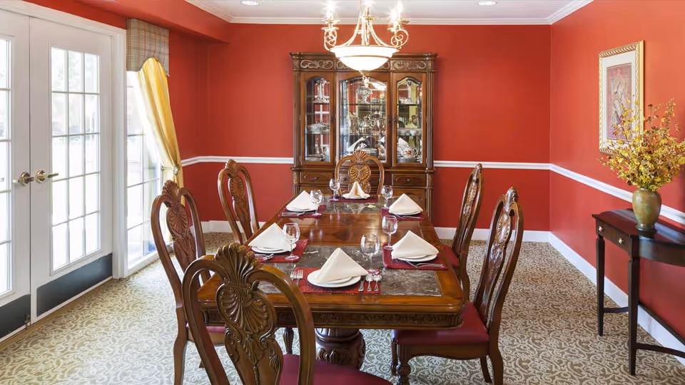 A formal dining room with red walls and white trim, featuring a wooden dining table set for six with white folded napkins, wine glasses, and silverware. The room has a patterned carpet, a chandelier overhead, a wooden china cabinet filled with dishes, and a small side table with a vase of yellow flowers. French doors with yellow curtains allow natural light into the room.
