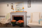 A cozy interior scene featuring a white fireplace mantel decorated with small pumpkins and autumn-themed items. Two cream-colored armchairs with light blue and green throws are positioned on either side of the fireplace. Two pots of orange chrysanthemums are placed in front of the fireplace. The walls are adorned with framed artwork.