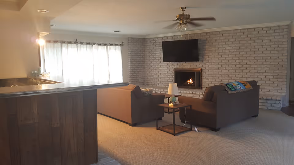 A cozy living room with two brown sofas facing a brick wall with a fireplace and a mounted flat-screen TV above it. A small side table with a lamp is placed between the sofas. To the left, there is a wooden counter and large windows with sheer curtains allowing natural light to fill the room.