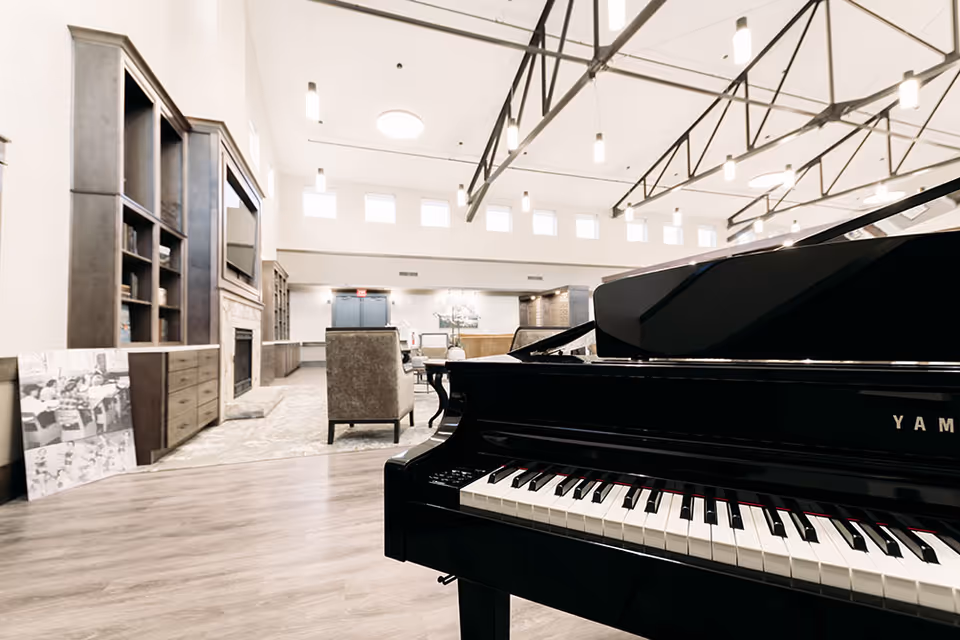 Spacious senior living common room with a glossy grand piano in the foreground, seating area, built-in shelving and high beamed ceilings.