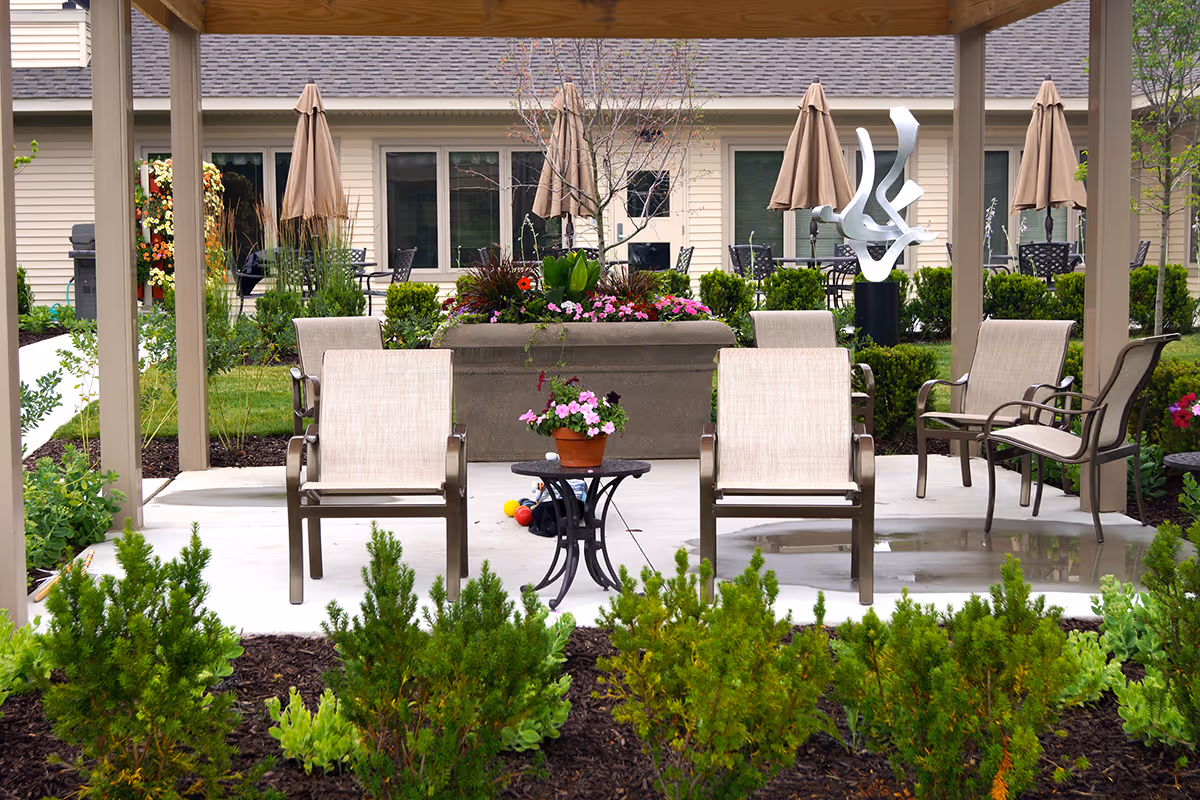 Outdoor patio area at Appledorn Assisted Living Center South featuring several beige cushioned chairs arranged around a small black table with a potted plant. The patio is covered by a pergola and surrounded by green shrubs and flower beds. In the background, there are beige buildings with windows, closed beige umbrellas, and a modern abstract metal sculpture.