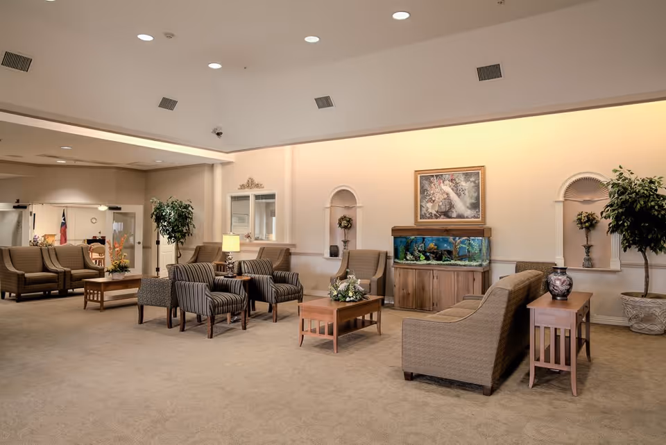 A spacious and well-lit living room area in a senior living facility with multiple seating arrangements including sofas and armchairs. The room features a large fish tank on a wooden stand, decorative plants, a painting on the wall, and side tables with lamps and floral arrangements. The walls are light-colored, and the ceiling has recessed lighting.