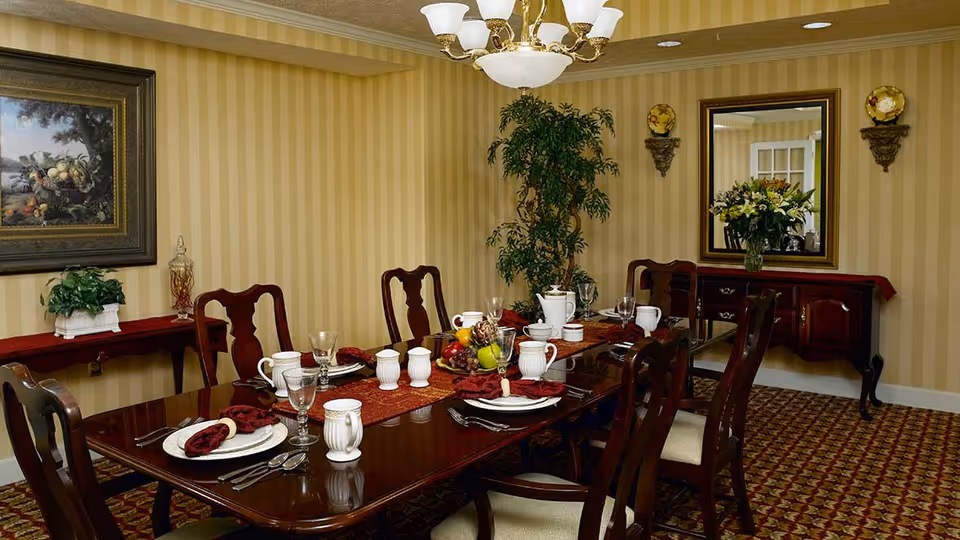 A formal dining room with a polished wooden table set for six people. The table has white plates, cups, silverware, and red cloth napkins. A centerpiece with fruit and pinecones is in the middle. The room features striped beige wallpaper, a large framed painting on one wall, a large mirror above a wooden sideboard with a flower arrangement, and a tall green plant in the corner.