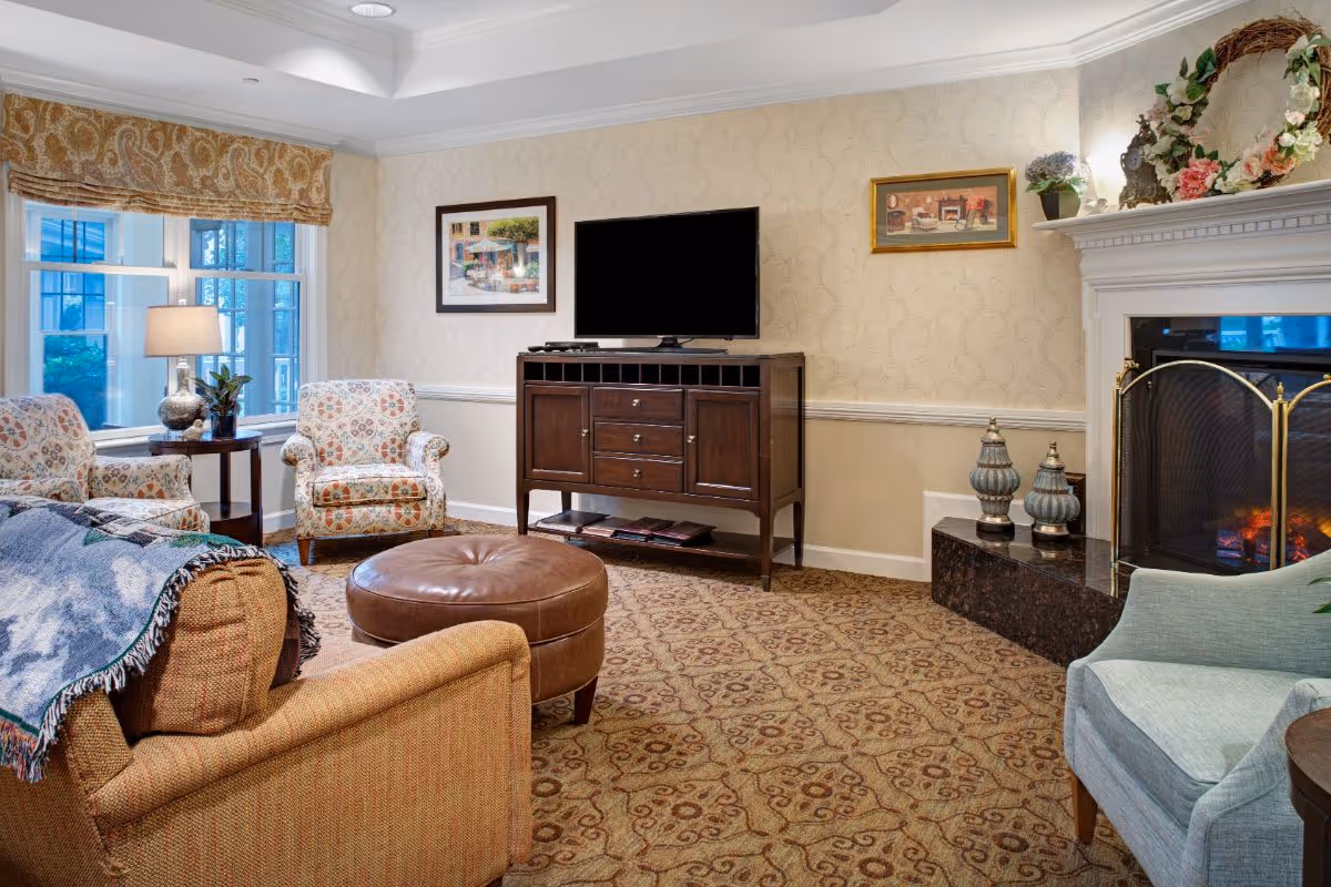 Cozy living room with upholstered chairs, a round leather ottoman, a TV on a wooden console, and a fireplace.