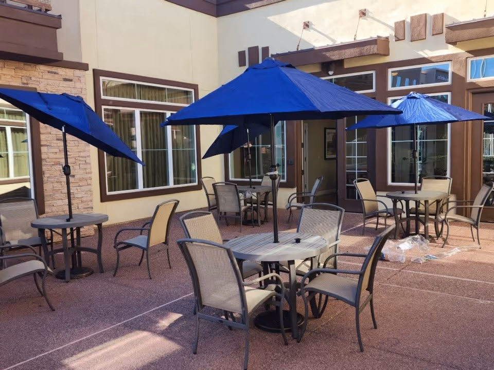 Outdoor patio area with multiple round tables, each surrounded by chairs and shaded by large blue umbrellas. The patio is adjacent to a building with large windows and doors.