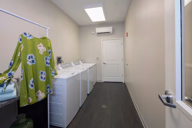 Narrow laundry room with three white top-loading machines along the left wall, a hanging green floral dress in the foreground, and a door at the far end.