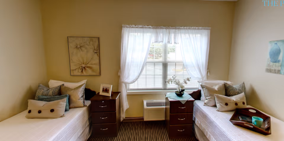 A cozy bedroom with two single beds placed on opposite sides of the room. Each bed has multiple pillows and a small wooden nightstand beside it. A window with sheer white curtains is centered on the back wall, allowing natural light to brighten the room. The walls are painted a soft beige color and decorated with framed artwork. A wooden tray with a cup and a book is placed on the right bed.