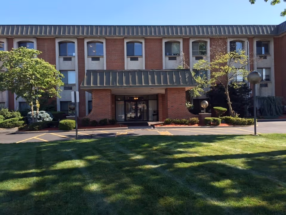 Front exterior view of a three-story brick and concrete building with a green metal awning over the main entrance. There are several windows on each floor, landscaped bushes and trees around the entrance, and a well-maintained lawn in the foreground.
