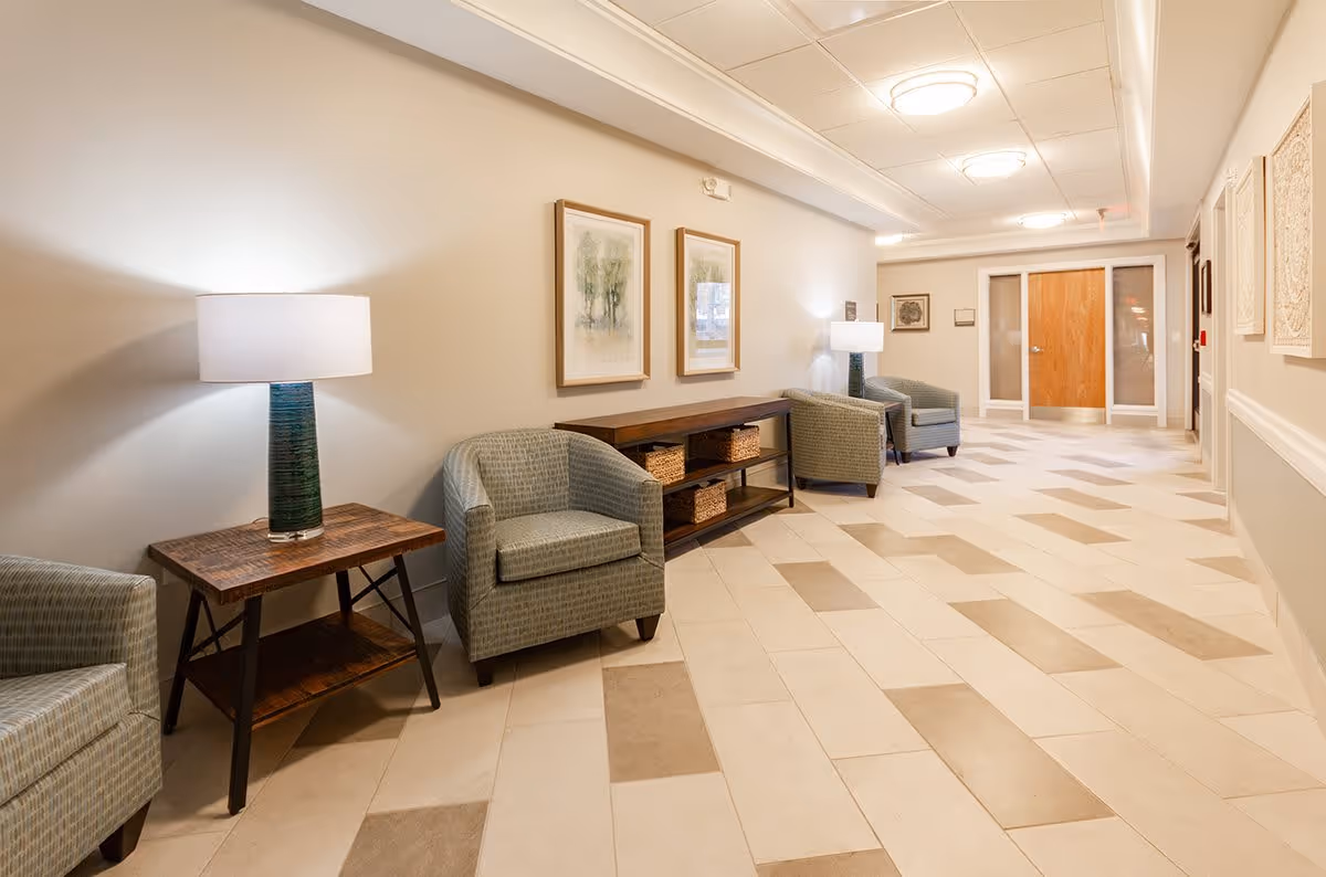 A well-lit hallway in a senior living facility with beige walls and tiled floor in a pattern of light and dark beige rectangles. The hallway features several cushioned armchairs, wooden side tables with lamps, framed artwork on the walls, and a wooden console table with baskets underneath. At the end of the hallway, there are double doors with glass panels.