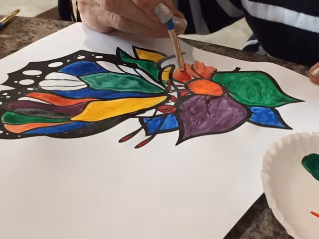 A close-up of a person painting a colorful butterfly design on a white sheet of paper using a small paintbrush. The butterfly wings are filled with vibrant colors including green, yellow, blue, purple, and red. The person is wearing a black and white striped shirt.