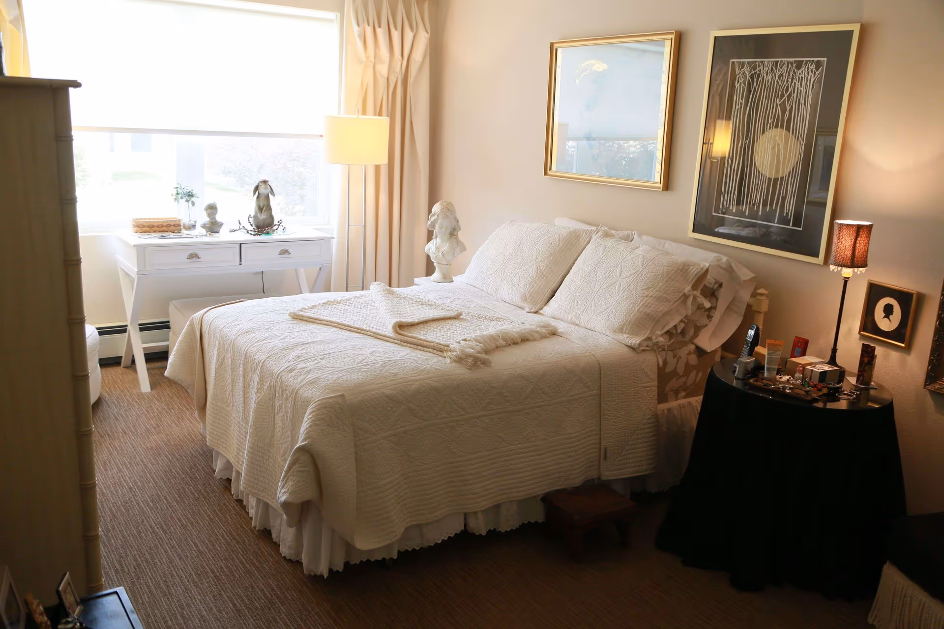 A cozy bedroom with a neatly made bed covered in white quilted bedding and pillows. There is a small white desk with decorative items and a lamp near a window with a white blind. On the right side of the bed, there is a round black table with various personal items and a small lamp. The walls have framed artwork and the floor is carpeted.