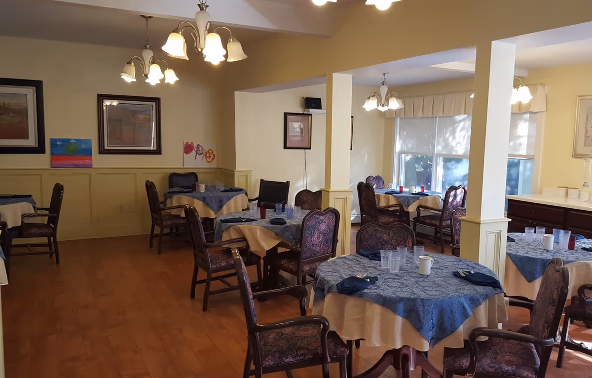 A dining room with several round tables covered with blue and beige tablecloths, each set with cups, glasses, and napkins. The room has wooden floors, yellow walls with framed artwork, and multiple chandeliers providing warm lighting. There are upholstered chairs around each table and large windows letting in natural light.