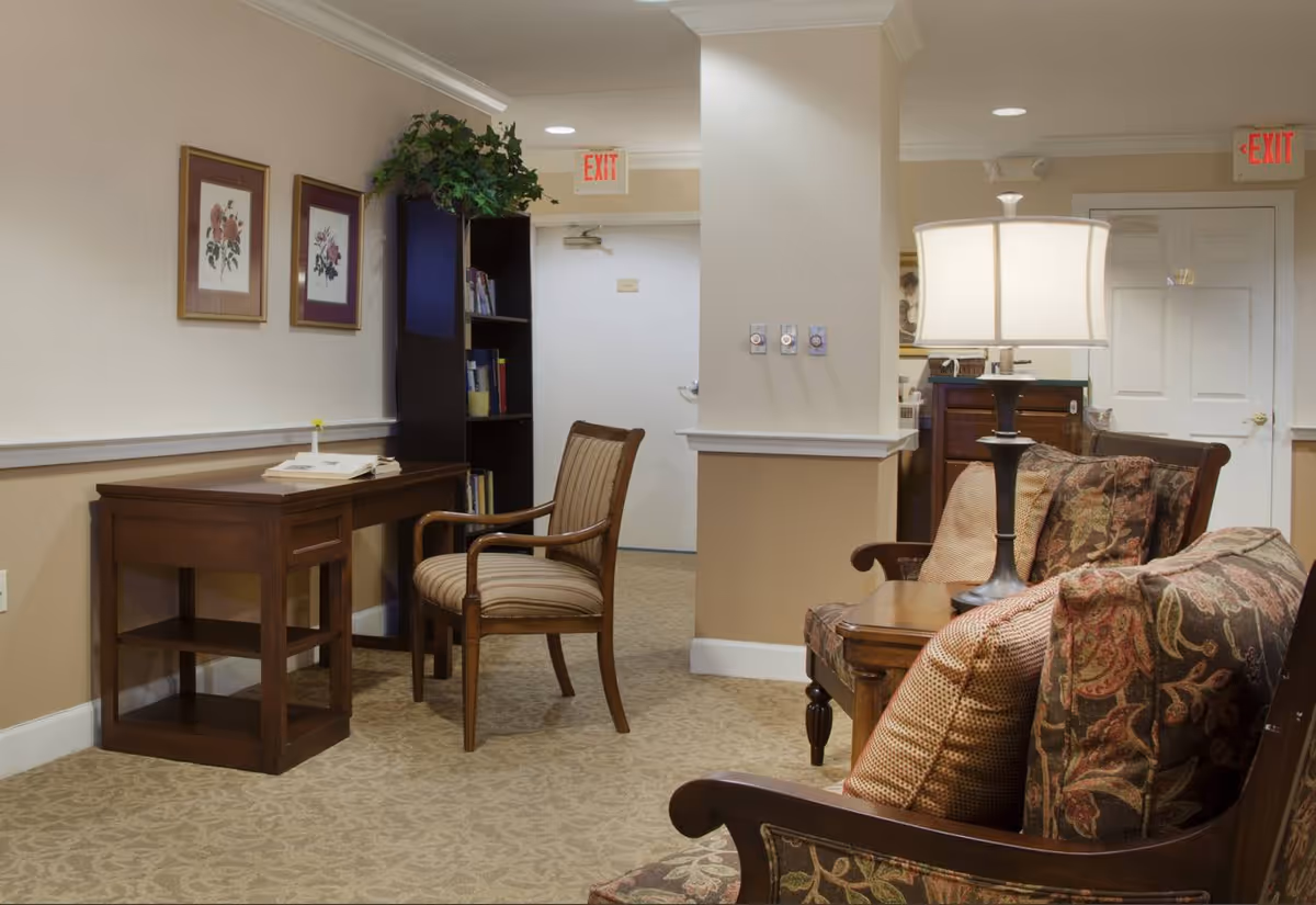 A cozy interior common area with a wooden desk and chair on the left, a tall bookshelf with books and a plant on top, and a seating area on the right featuring a cushioned armchair and sofa with patterned pillows. A table lamp is placed on a wooden side table between the sofa and armchair. The walls are painted beige with white trim, and there are two framed floral pictures hanging above the desk. Two exit signs are visible above doors in the background.