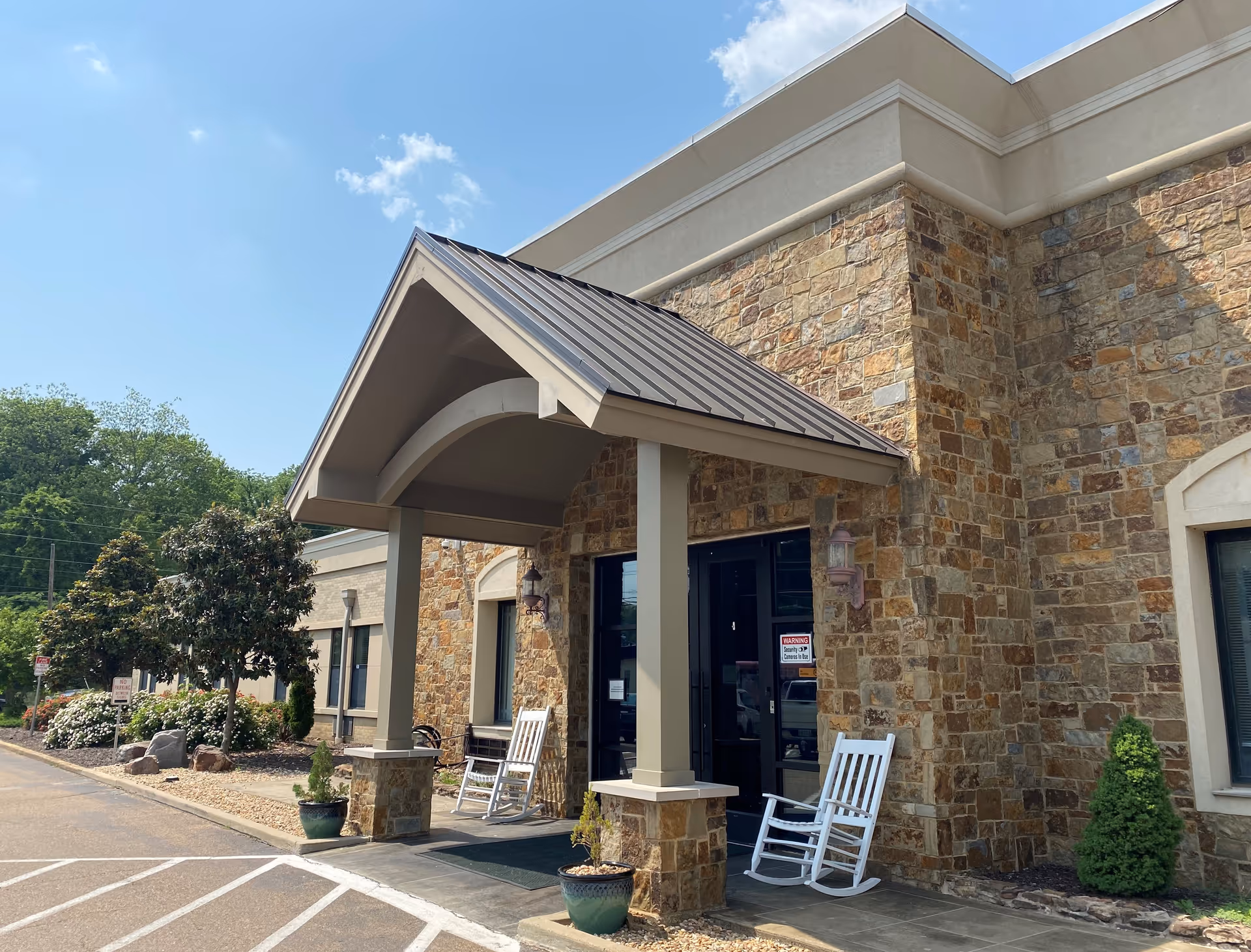 Entrance of a building with stone exterior walls and a covered porch supported by columns. Two white rocking chairs are placed on the porch near the glass double doors. There are potted plants on either side of the entrance and landscaping with bushes and trees along the side of the building. The sky is clear with a few clouds.