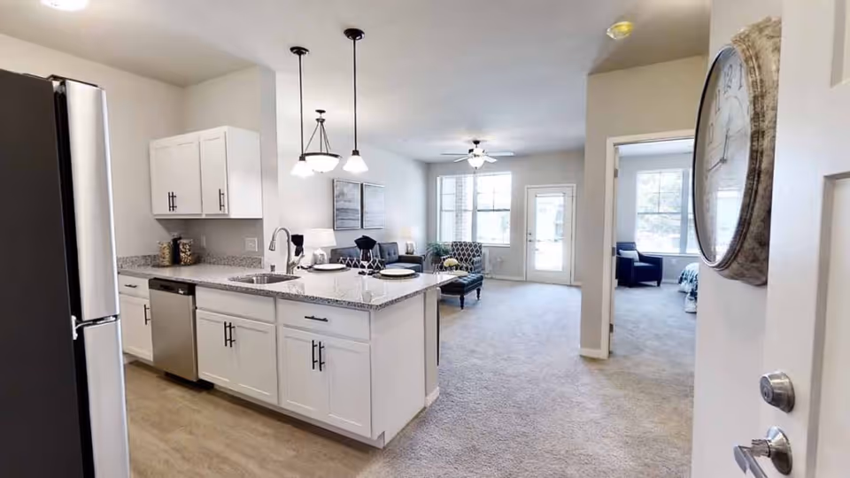 View of a modern open-concept apartment interior at Aspire Oshkosh, featuring a kitchen with white cabinets, granite countertops, stainless steel refrigerator and dishwasher, pendant lights above the kitchen island, and a living area with carpet flooring, a sofa, and a large window letting in natural light. A doorway leads to a bedroom with a chair and bed visible. A large wall clock is mounted near the entrance door.