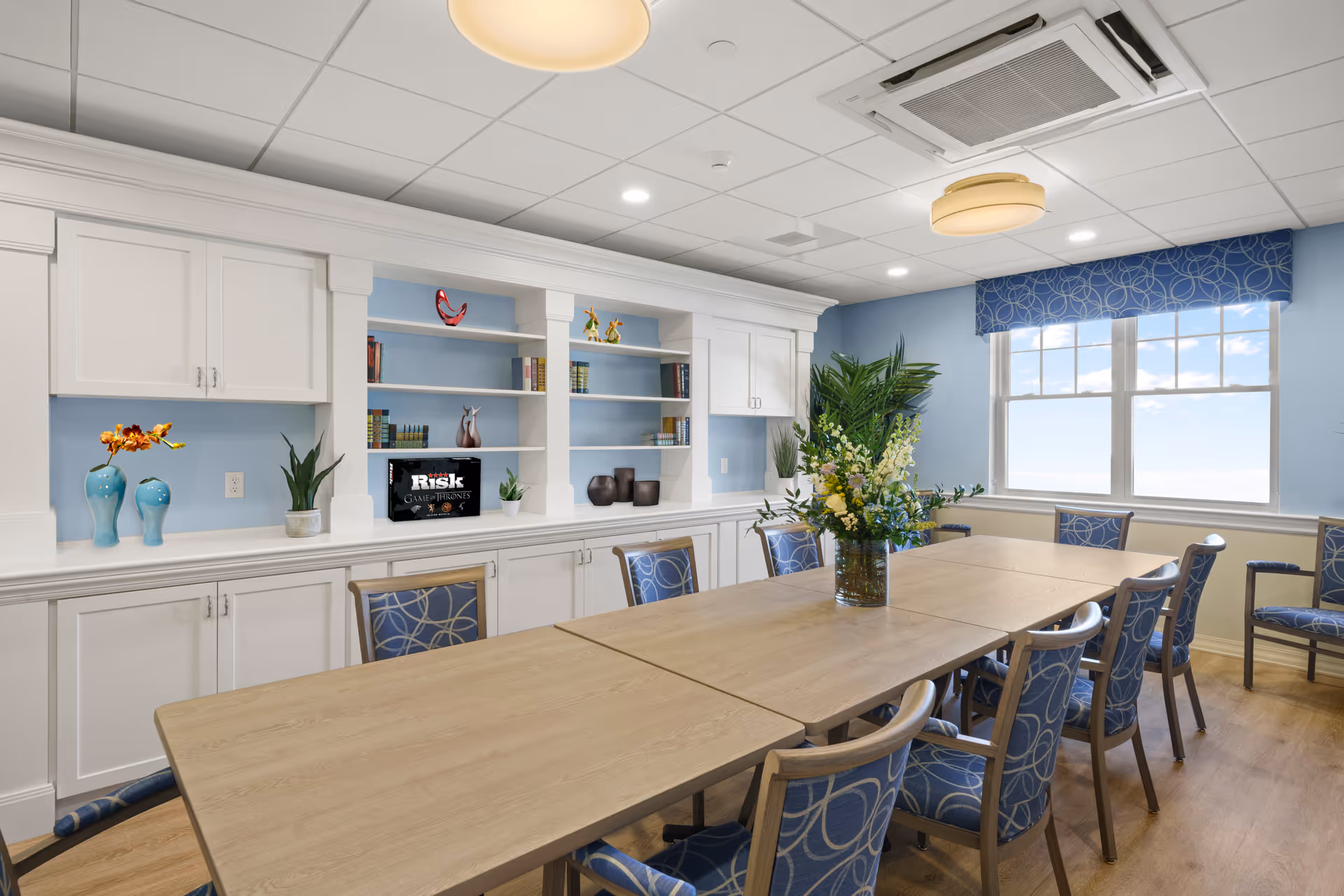 A bright and clean meeting or activity room with a long wooden table surrounded by blue patterned chairs. The room has light blue walls, white built-in cabinets and shelves with books and decorative items, a large window with a blue valance letting in natural light, and a vase with flowers on the table.
