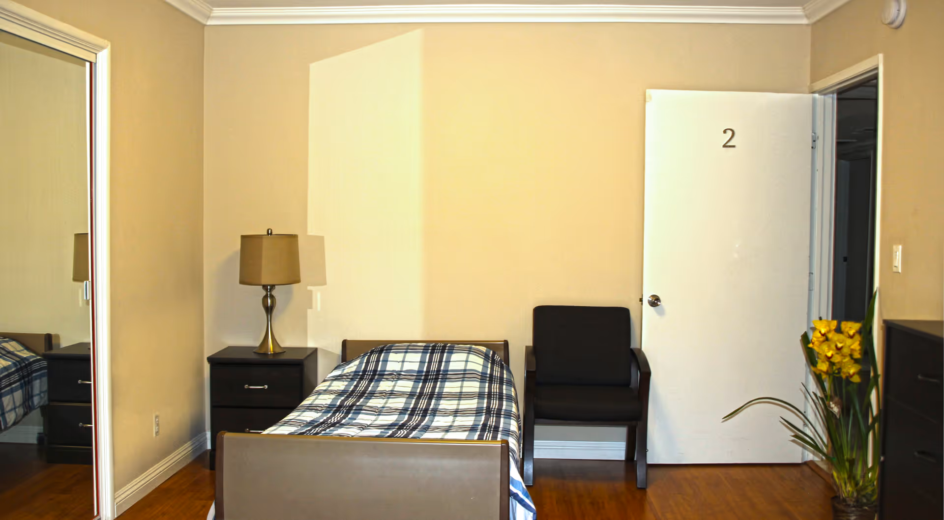 A simple senior living bedroom with a single bed covered in a plaid blanket, a nightstand with a lamp, a black armchair, a white door labeled with the number 2, a large mirror on the left wall, and a plant with yellow flowers on the right side.