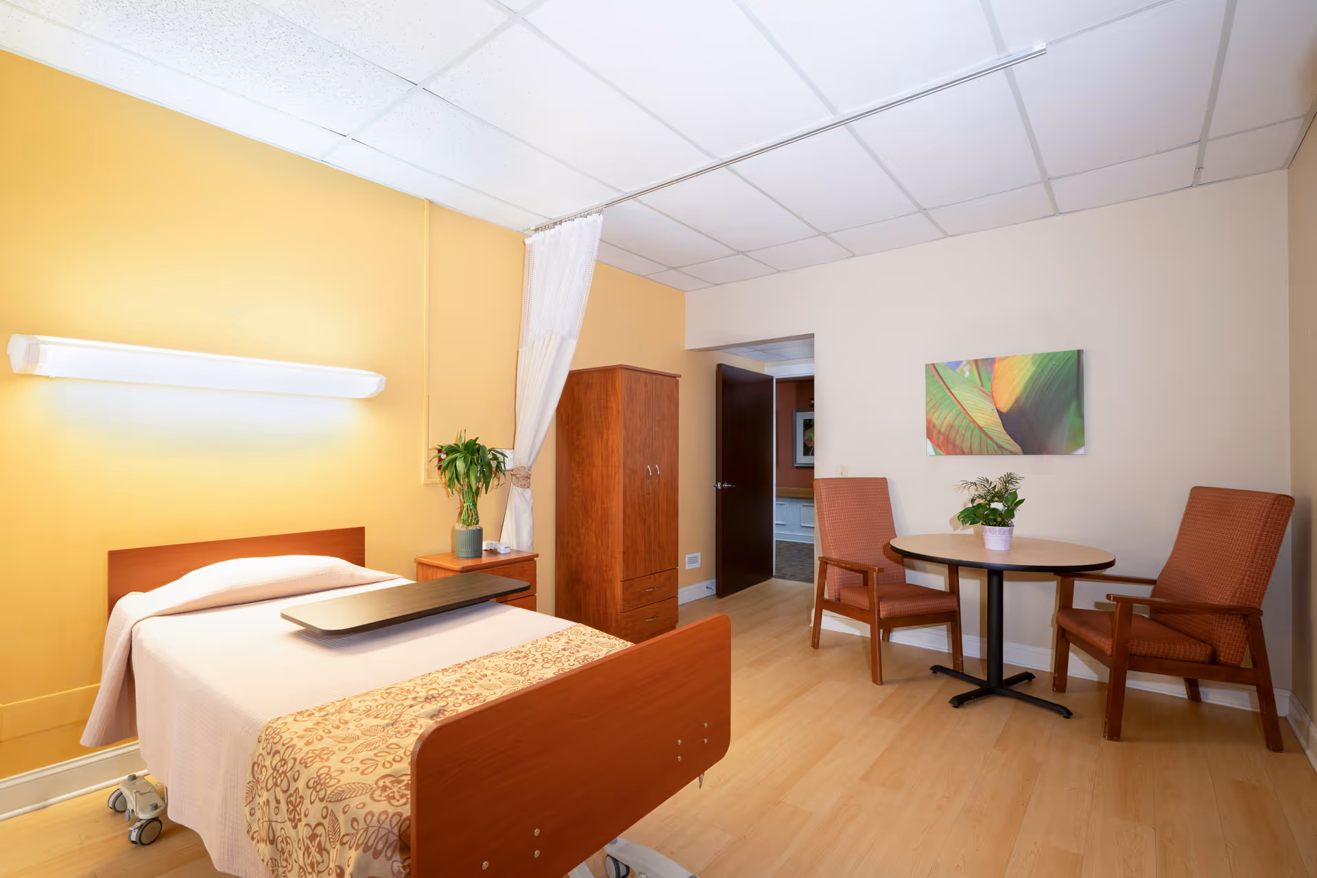 A well-lit senior living bedroom with a hospital-style bed covered with a beige blanket and a floral-patterned throw. Next to the bed is a small wooden nightstand with a potted plant. A wooden wardrobe stands against a yellow wall. On the opposite side, there is a small round table with two orange cushioned chairs and a potted plant on the table. A colorful leaf-themed painting hangs on the beige wall above the table. The room has a light wood floor and a white drop ceiling with fluorescent lighting.