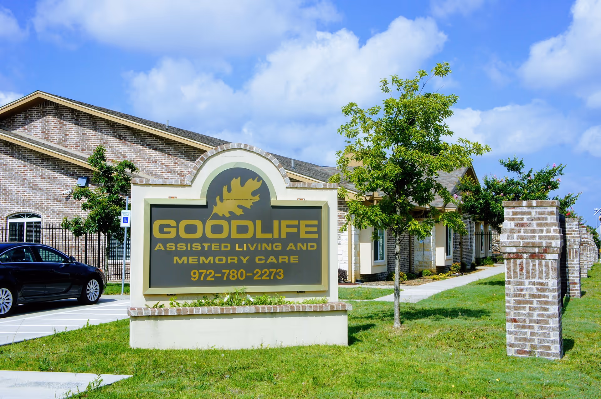 Outdoor view of the Goodlife Assisted Living and Memory Care facility sign with a brick building and green lawn under a blue sky with clouds.