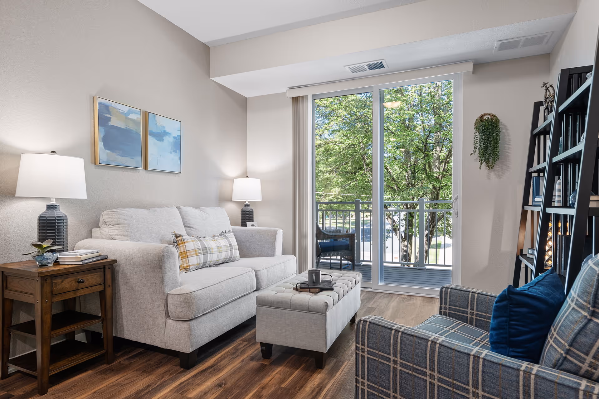 A cozy living room with a light gray sofa adorned with a plaid pillow, a matching ottoman with a tray and cup on it, a wooden side table with a lamp and books, and a plaid armchair with a blue pillow. The room has wood flooring, two abstract paintings on the wall, a tall black bookshelf filled with books and decor, and a large sliding glass door leading to a balcony with outdoor seating and green trees visible outside.