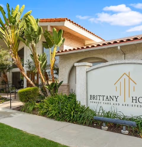 Exterior view of Brittany House Residential Memory Care facility showing a beige stucco building with a tiled roof, surrounded by green plants and palm trees, with a large sign displaying the facility's name.
