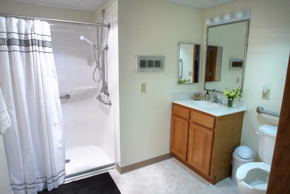 A clean bathroom featuring a walk-in shower with a white curtain and grab bars, a wooden vanity with a sink and mirror above it, a toilet with a grab bar beside it, and a small trash can. The walls are light-colored and the floor is tiled.