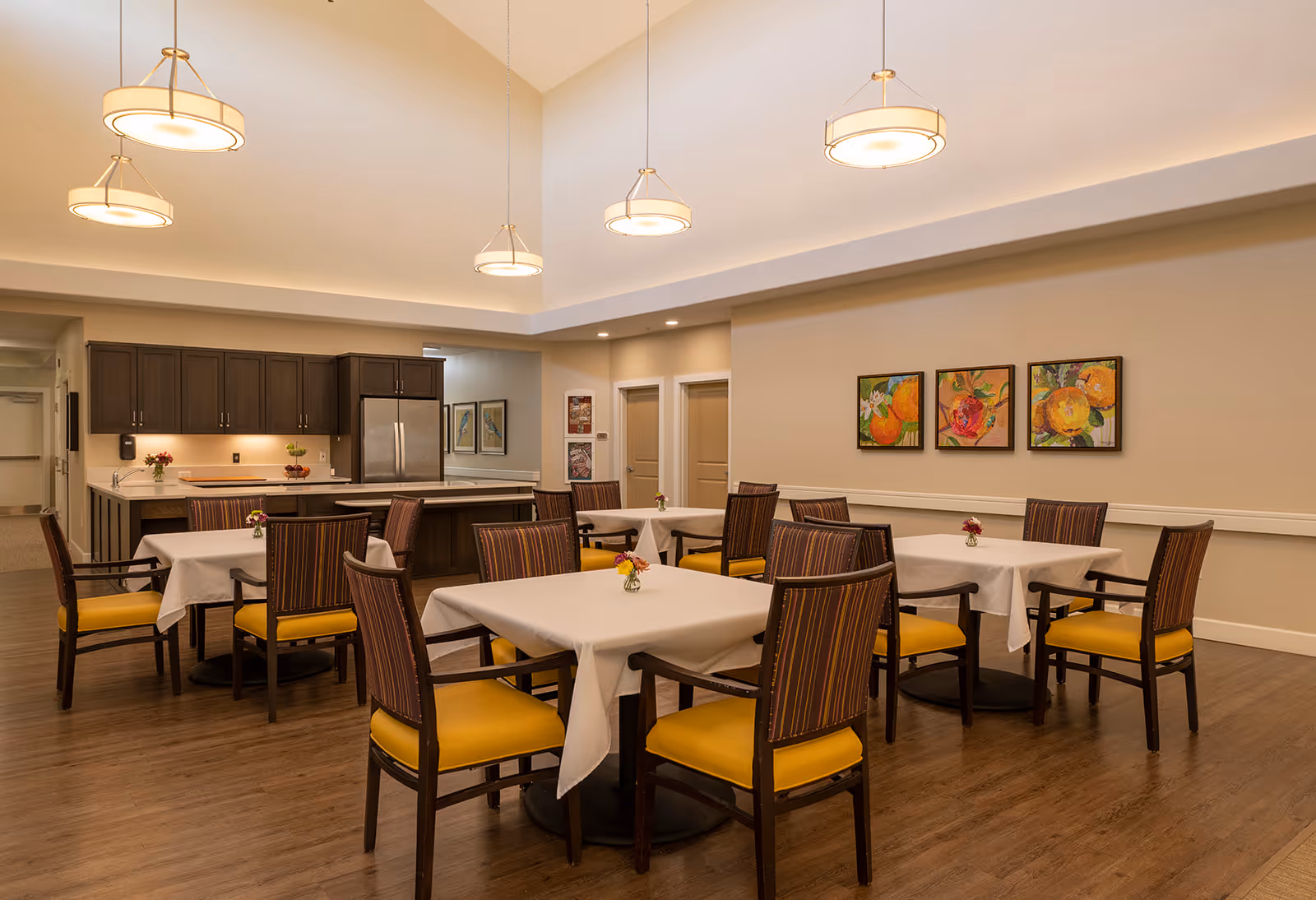 A dining room with several tables covered in white tablecloths, each surrounded by chairs with yellow seats and striped backs. The room has a high ceiling with modern hanging light fixtures. In the background, there is a kitchen area with dark wood cabinets and a stainless steel refrigerator. Three colorful floral paintings hang on the wall.