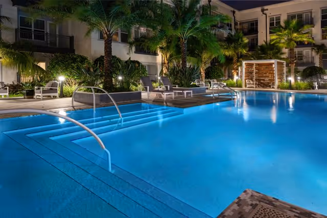 Night view of a well-lit outdoor swimming pool surrounded by palm trees and lounge chairs, with a modern building in the background and a wooden pergola seating area on the right side.