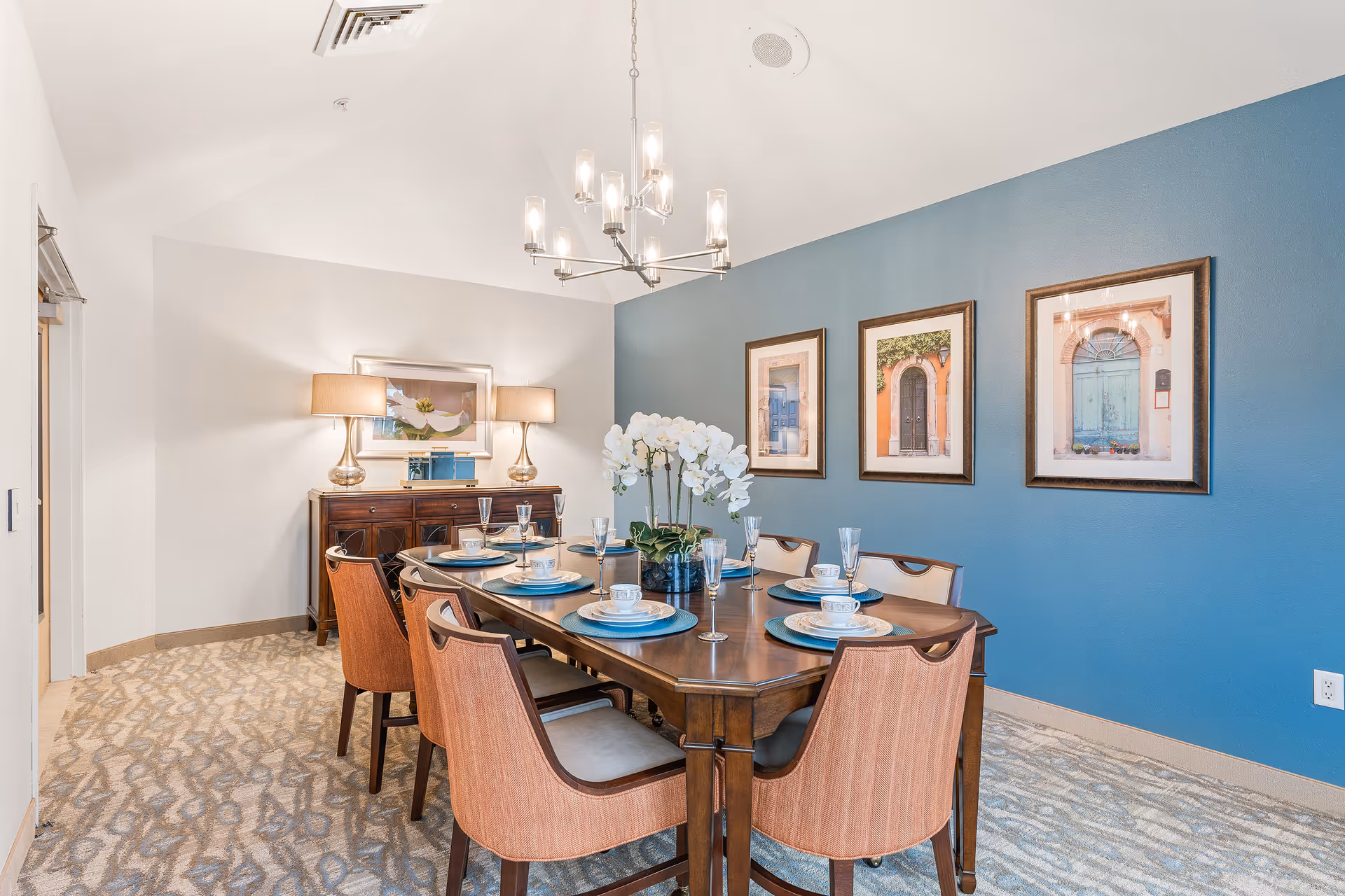 Formal dining room with a wooden table set for six, a blue accent wall with framed artwork, a chandelier, a sideboard with lamps, and an orchid centerpiece.