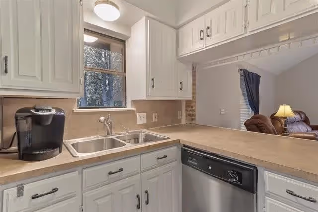 Kitchen with a double sink, dishwasher, white cabinets, countertop and a coffee maker, open to a living area.