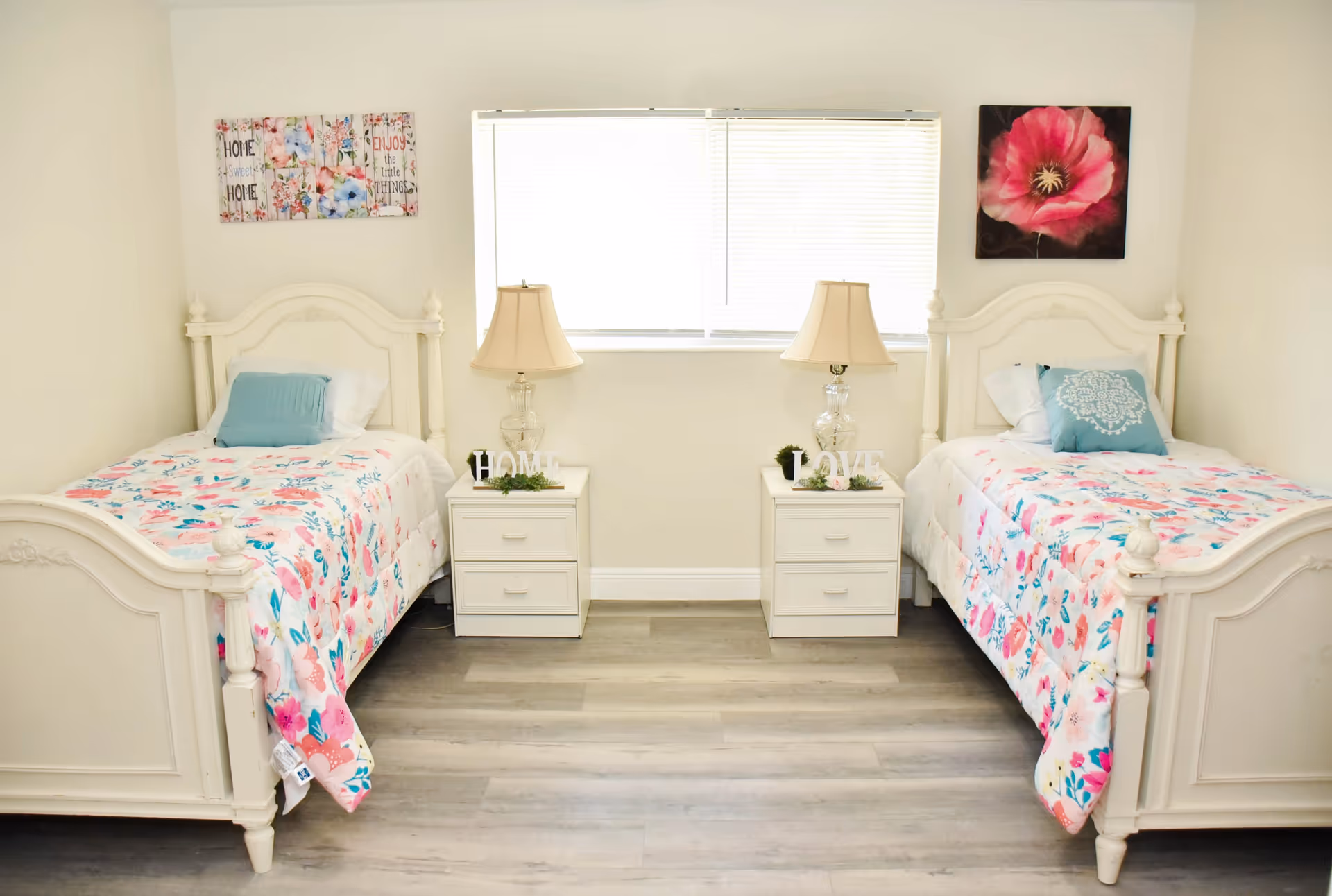 A bright bedroom with two white twin beds featuring floral bedspreads and blue accent pillows. Between the beds are two white nightstands, each with a clear glass lamp and decorative items spelling 'HOME' and 'LOVE'. Above each bed hangs a floral-themed wall art. A window with blinds is centered on the wall behind the nightstands.