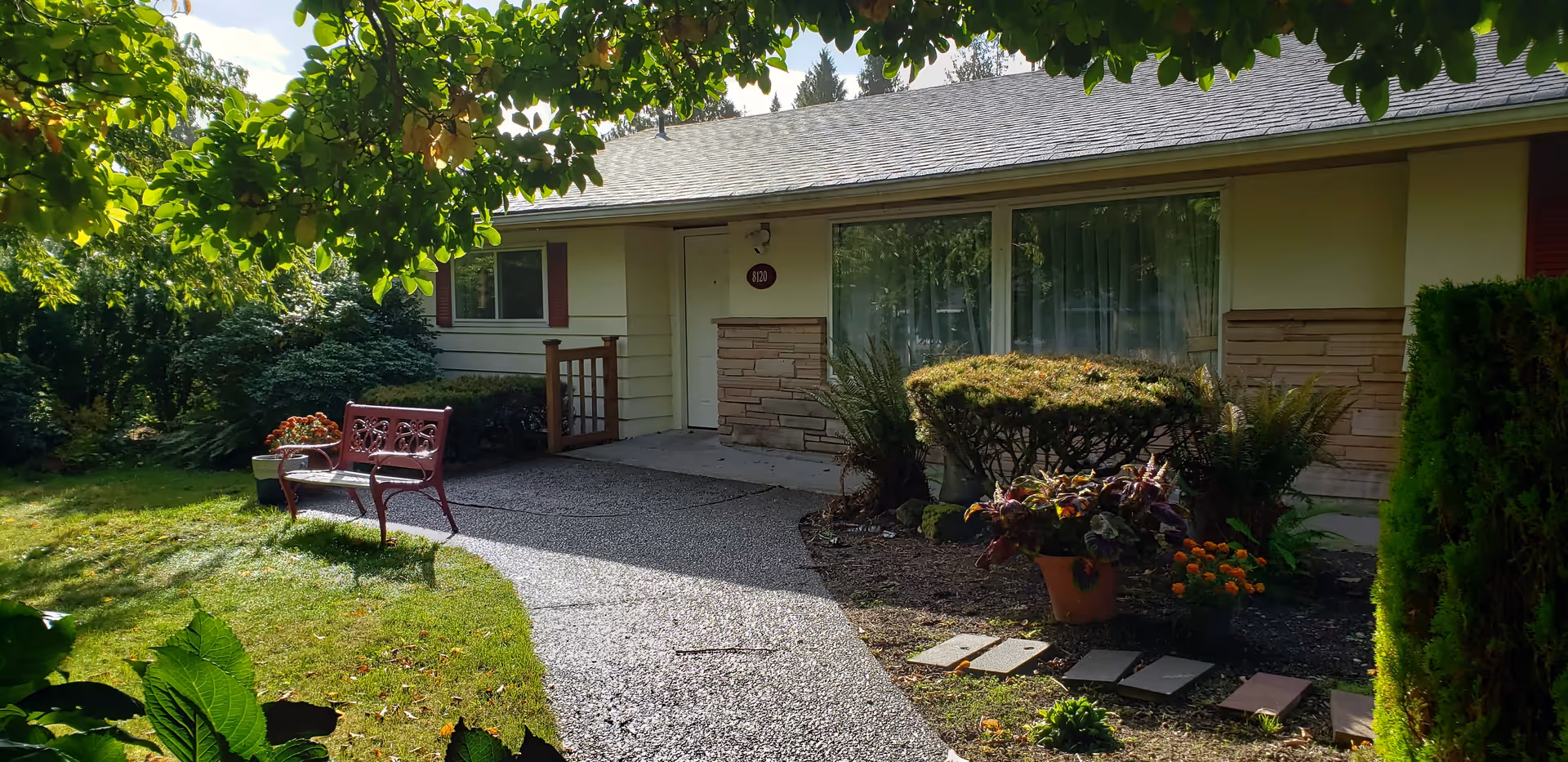 Front exterior view of a single-story house with a pathway leading to the entrance. There is a red bench on the left side of the path, surrounded by green bushes, plants, and a small garden area. The house has light-colored siding with stone accents and large windows with curtains.