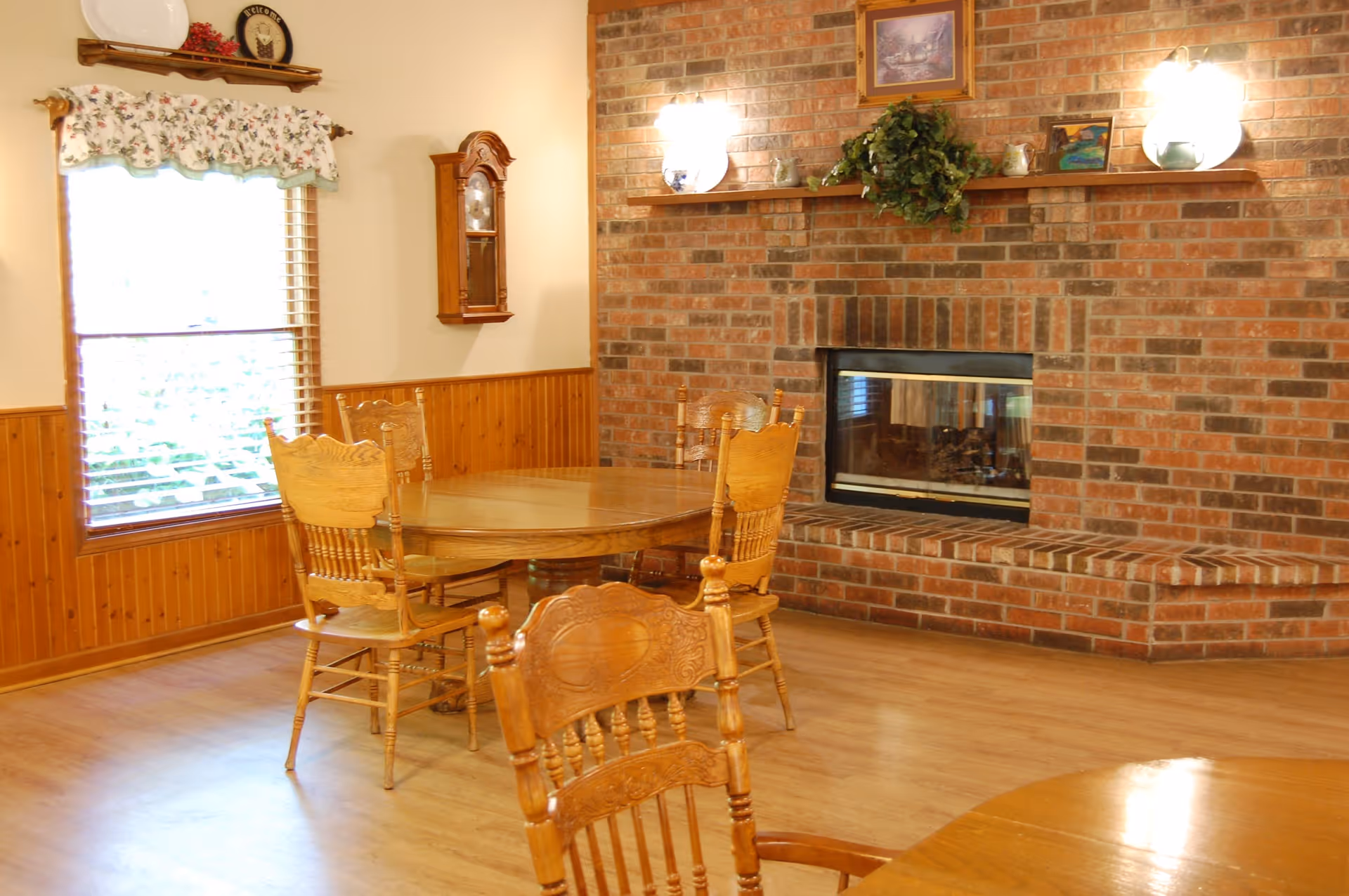 A cozy dining area with wooden chairs and tables, a large window with floral curtains, and a brick fireplace with a wooden mantel decorated with plants and framed pictures.