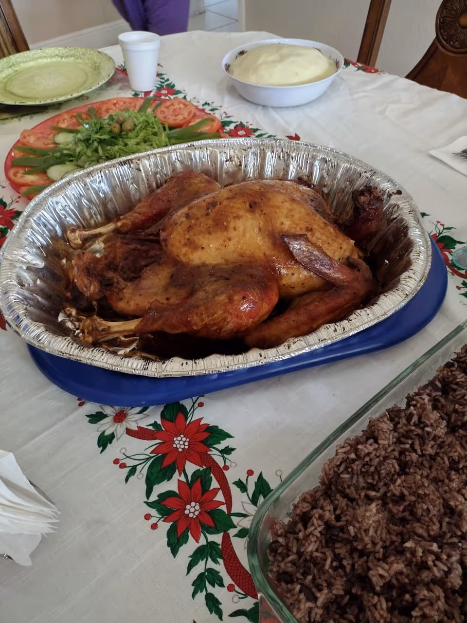 A table set with a roasted whole chicken in an aluminum tray, a glass dish of rice and beans, a bowl of mashed potatoes, a plate with sliced tomatoes, cucumbers, lettuce, and olives, and a green plate with a white cup in the background. The tablecloth has a festive red and green floral pattern.